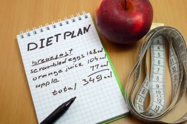 Notebook with diet plan, apple, and measuring tape on table.